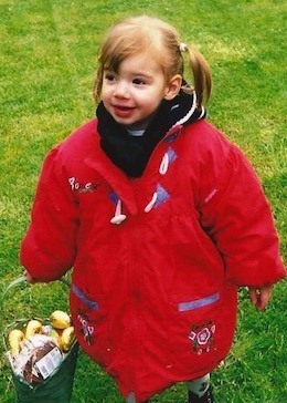 A young Carla wearing an oversized red coat and carrying a green basket