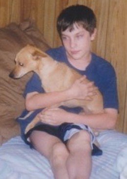 Un jeune Patrick en chandail bleu et short noir assis sur son lit tenant un chien