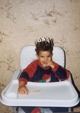 A young Adil in his pajamas wearing a foil crown and eating in a high chair
