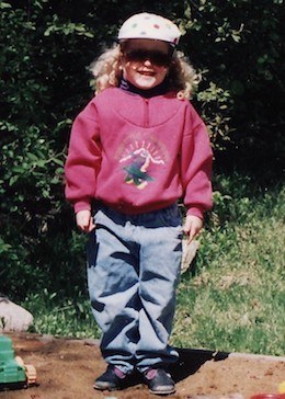 A young Alyssa Therrien-Coulombe wearing jeans and a pink sweatshirt, standing in a sandbox