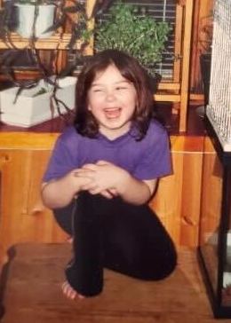 A young Marili wearing a purple shirt and black pants, sitting on a table and laughing