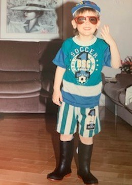 A young Mathieu wearing a hat, sunglasses, a soccer t-shirt, striped shorts, and rain boots